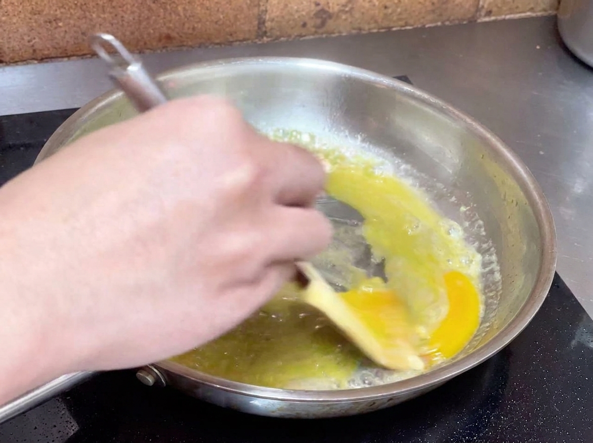 A hand vigorously stirring a yellow oil and water mixture in a pan with a spatula to create a creamy sauce.