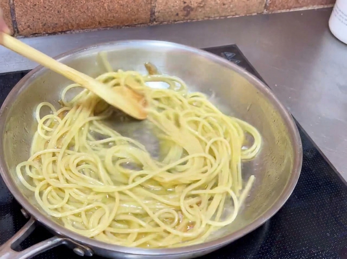 Spaghetti being tossed and stirred in a pan with a wooden spoon to coat the noodles in garlic oil sauce.