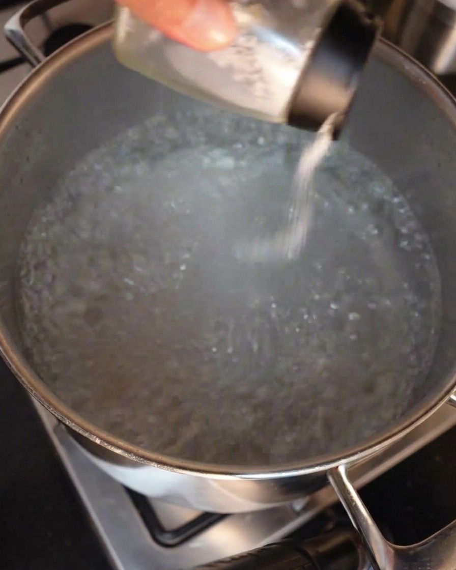 A hand sprinkling salt from a glass shaker into a large stainless steel pot filled with actively boiling water.