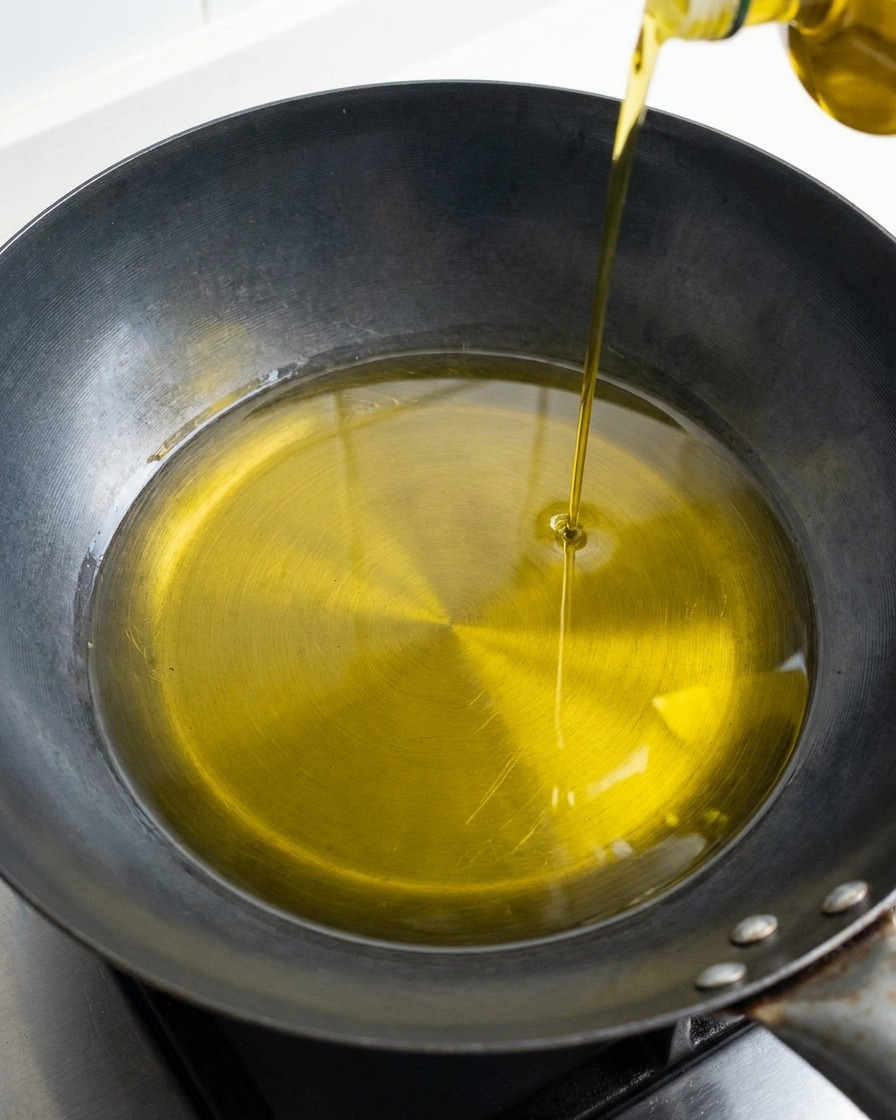 A steady stream of golden olive oil being poured from a bottle into the center of a dark, dry wok or pan.