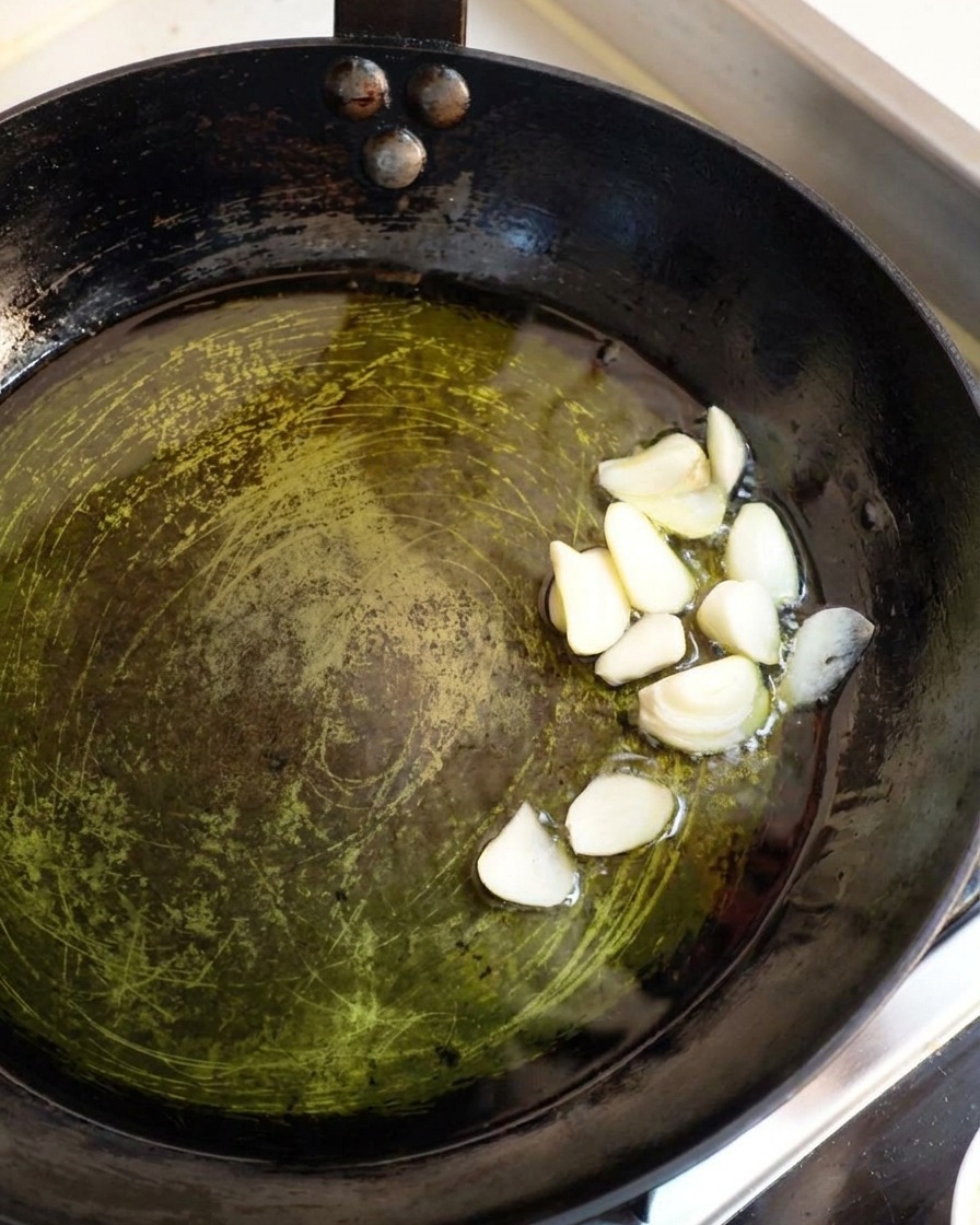 Thick slices of raw white garlic simmering gently in a shallow pool of hot olive oil inside a dark pan.