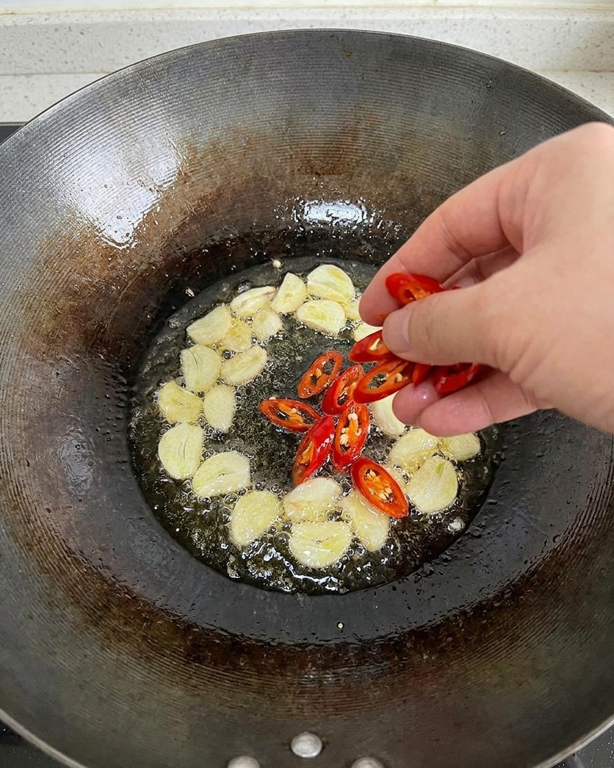 A hand dropping freshly sliced red chili peppers into a wok of hot oil that is already bubbling with garlic slices.