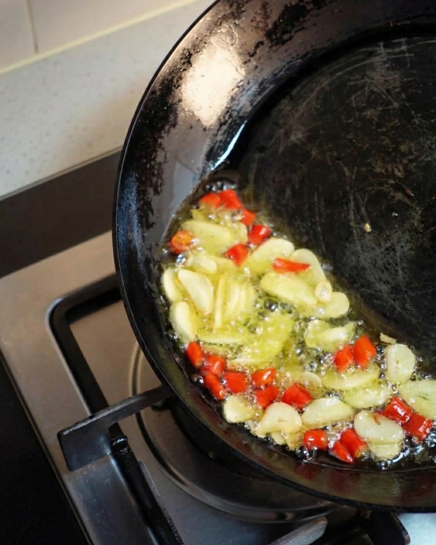 A dark wok tilted to one side, pooling hot golden oil where sliced garlic and chopped red chilies are vigorously bubbling and frying together.