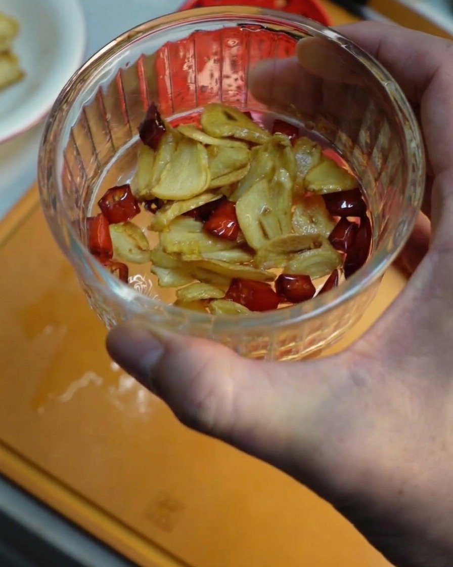 A hand holding a small glass bowl filled with crispy fried garlic slices and red chili pieces.