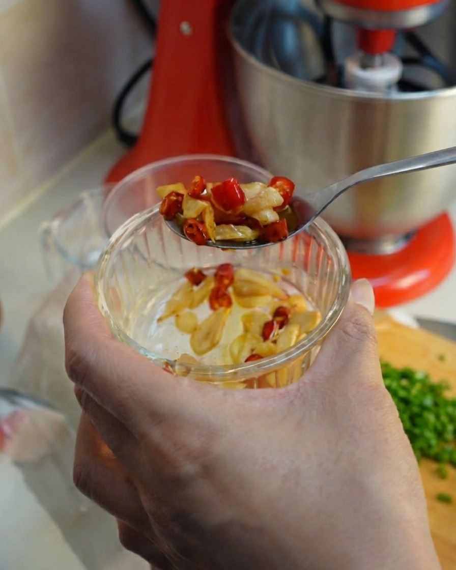 A spoon scooping up fried garlic and red chili slices from a small glass bowl.