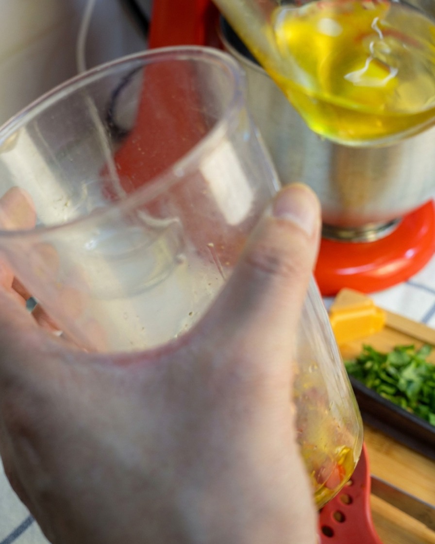 An immersion blender being lifted from a clear cup, dripping with a smooth, golden-red blended garlic and chili sauce.