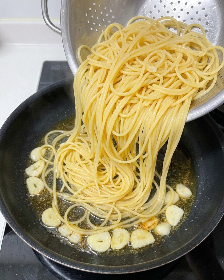 Cooked spaghetti being transferred from a metal colander into a skillet containing infused oil and fried garlic slices.