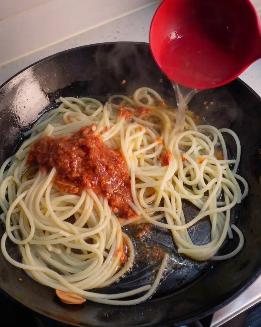 A red ladle pouring clear pasta water into a skillet over spaghetti and a spoonful of blended red garlic chili sauce.