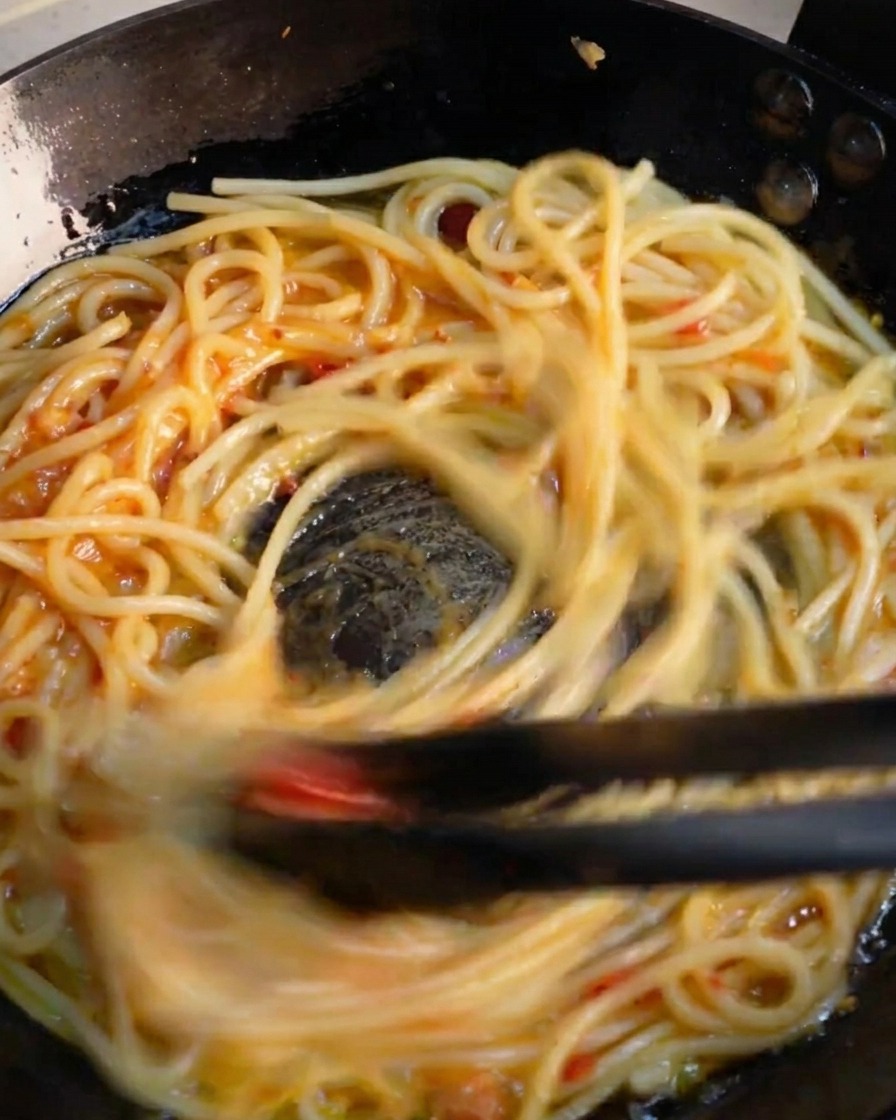 Black tongs rapidly tossing spaghetti in a skillet to emulsify the vibrant garlic and chili sauce.
