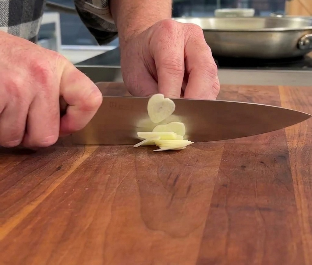 Hands using a chefs knife to thinly slice a clove of garlic on a wooden cutting board.