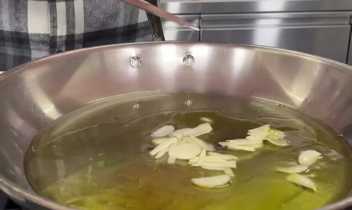 Using a knife blade to slide thinly sliced garlic into a stainless steel pan filled with warm olive oil.
