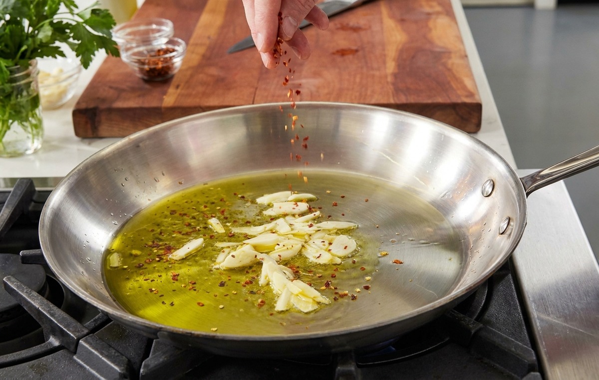 A hand sprinkling red pepper flakes into a pan of olive oil and sliced garlic.