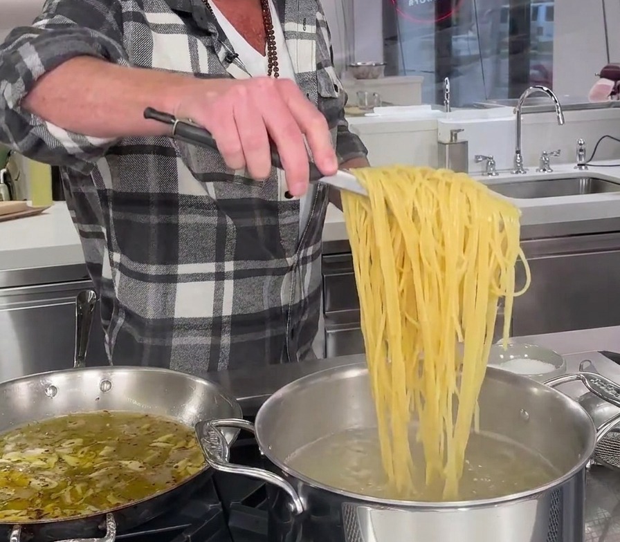 Partially cooked spaghetti being lifted straight from a pot of boiling water with long metal tongs.