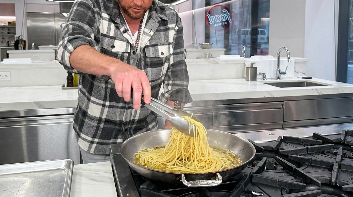 Partially cooked spaghetti being dropped into a hot pan with garlic, oil, and pasta water using metal tongs.
