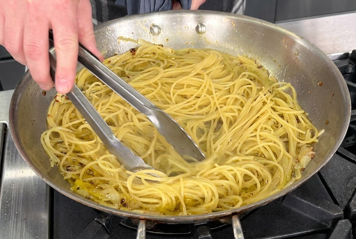 Using metal tongs to toss cooked spaghetti in a stainless steel pan with olive oil, garlic, and red pepper flakes.
