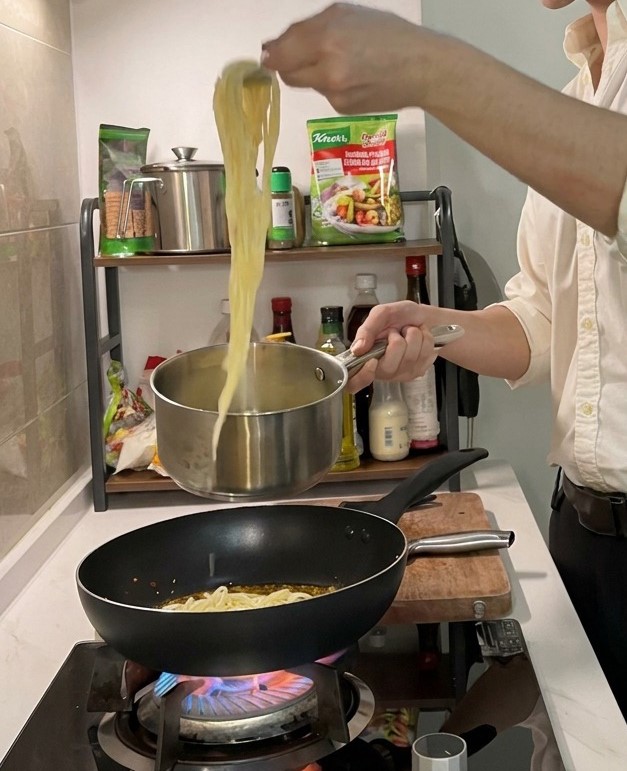 Long strands of cooked spaghetti being lifted from a stainless steel pot with tongs, ready to be added to a hot dark frying pan containing seasoned oil.