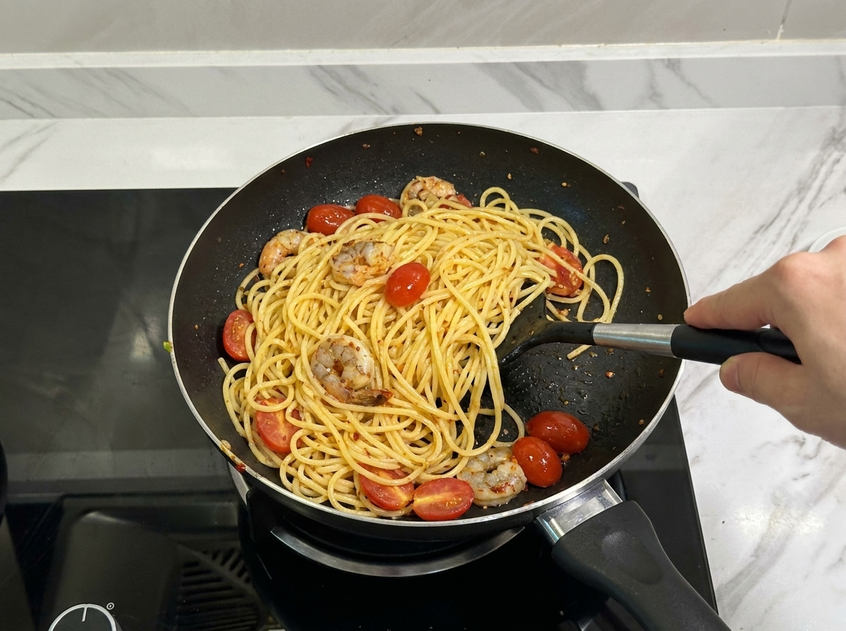 A black spatula tossing spaghetti, seasoned shrimp, and halved cherry tomatoes together in a frying pan.