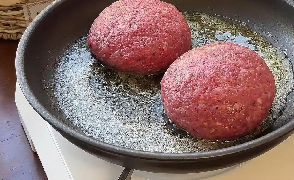 Two thick, raw beef burger patties being pan-fried in melted butter on a portable stove.