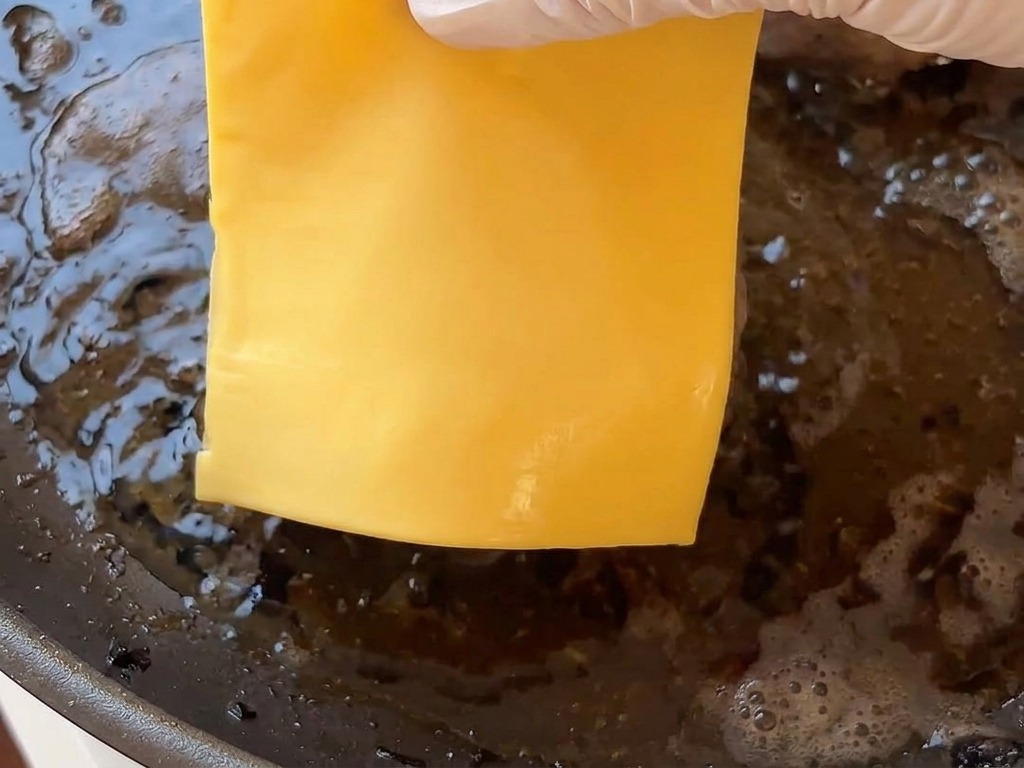 A gloved hand lowering a square of yellow cheese onto a hot burger patty cooking in a pan.