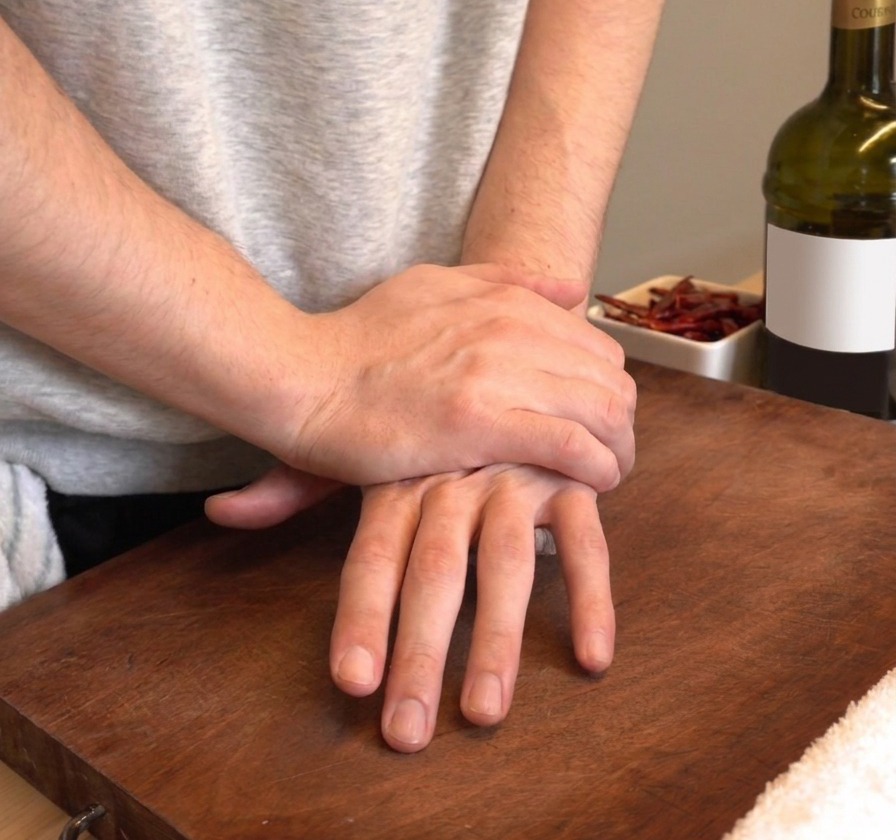 Hands pressing down firmly to smash fresh garlic cloves on a wooden cutting board.