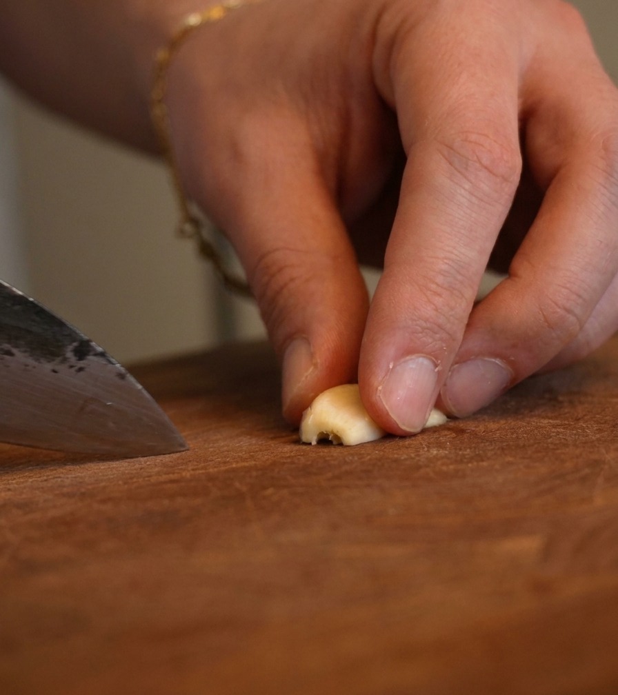 Close-up of fingers holding a piece of garlic while a chefs knife finely minces it.