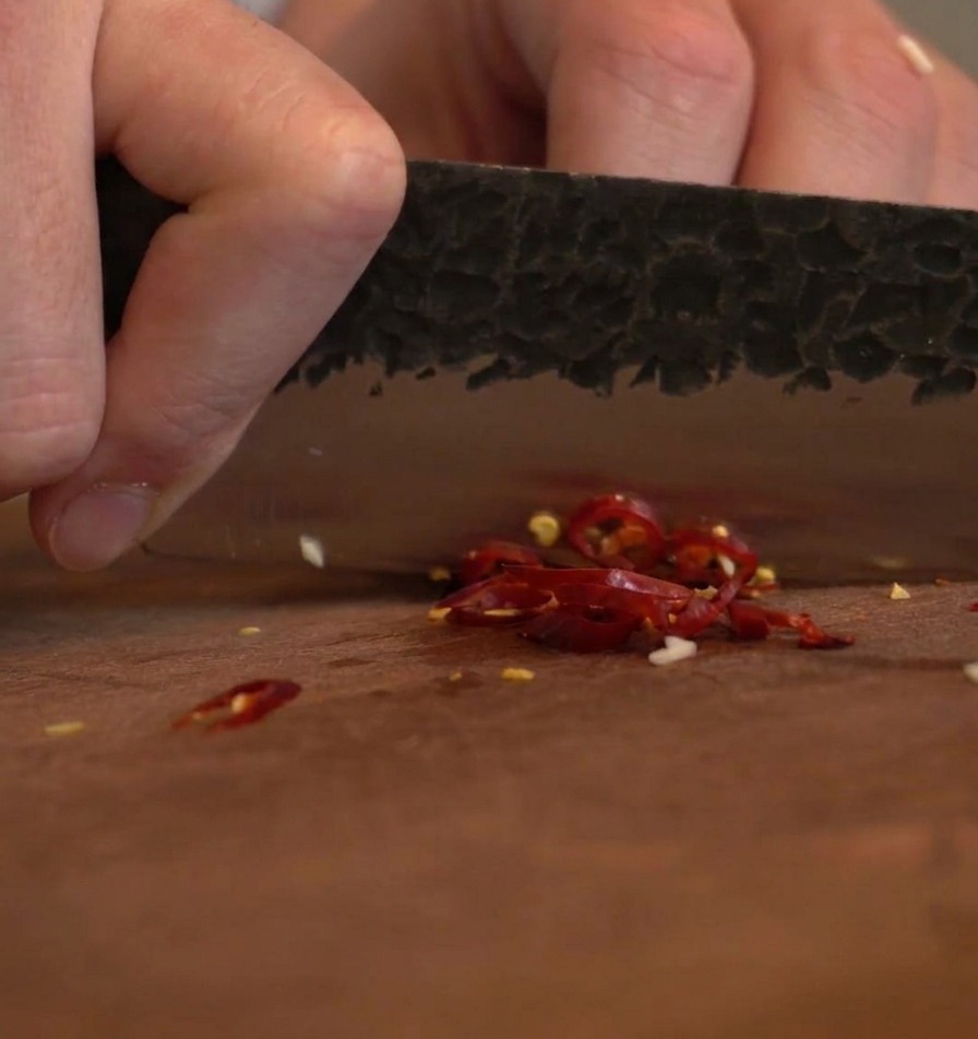 A textured dark knife blade chopping vibrant dry red chilies into pieces on a wooden board.