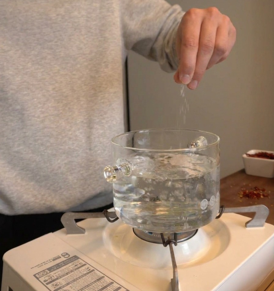 Hand sprinkling salt into a clear glass pot of bubbling boiling water over a portable stove.