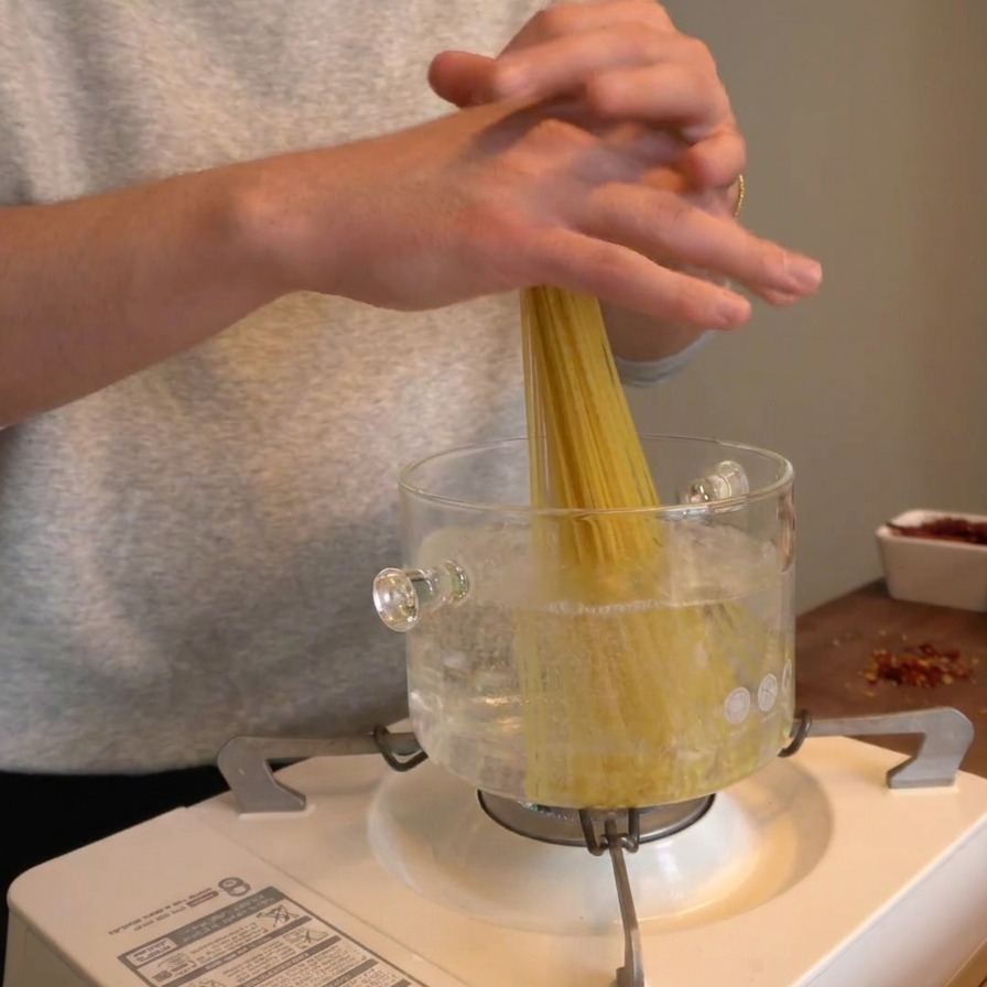 Hands dropping a bunch of dry spaghetti vertically into a clear glass pot of boiling water.