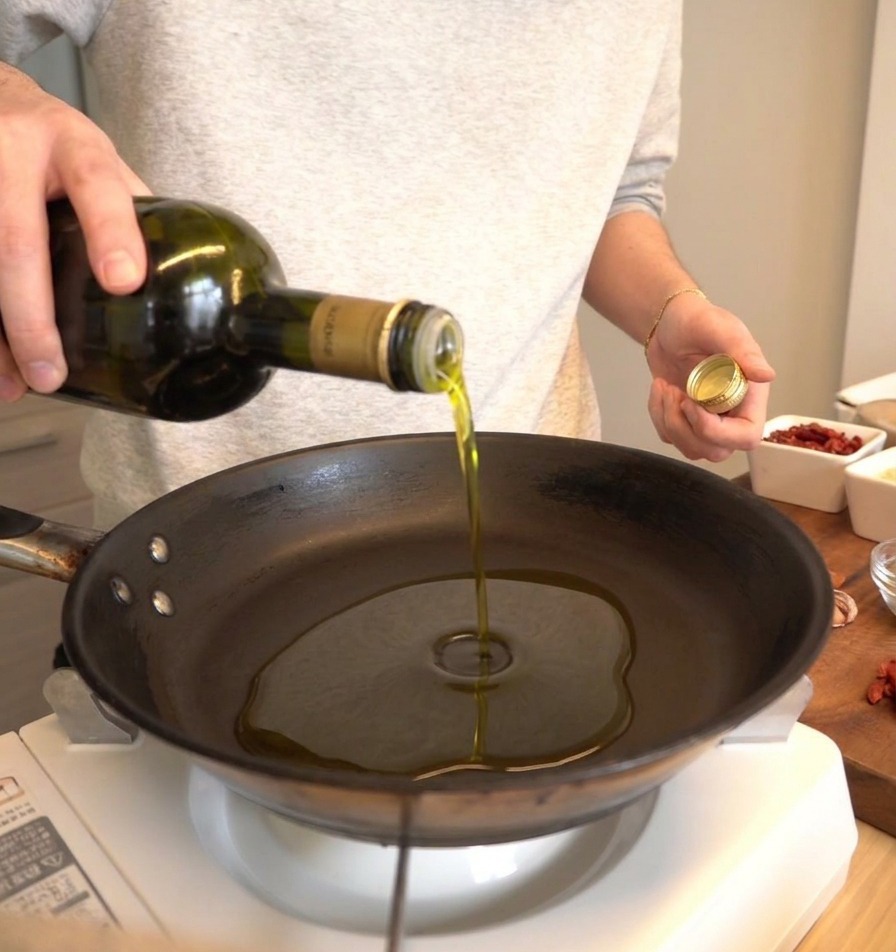 Olive oil being poured from a dark green bottle into a black skillet.