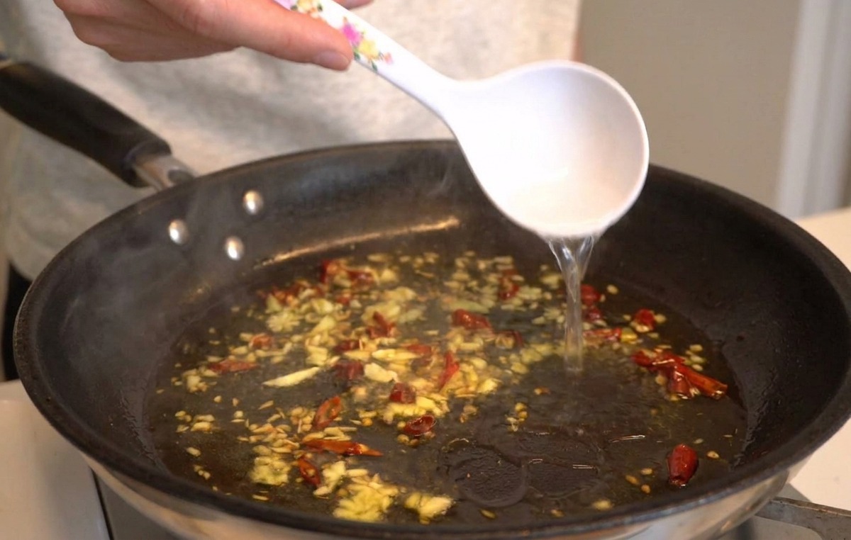 A white ladle pouring hot starchy water into a pan containing sizzling garlic, oil, and chilies.
