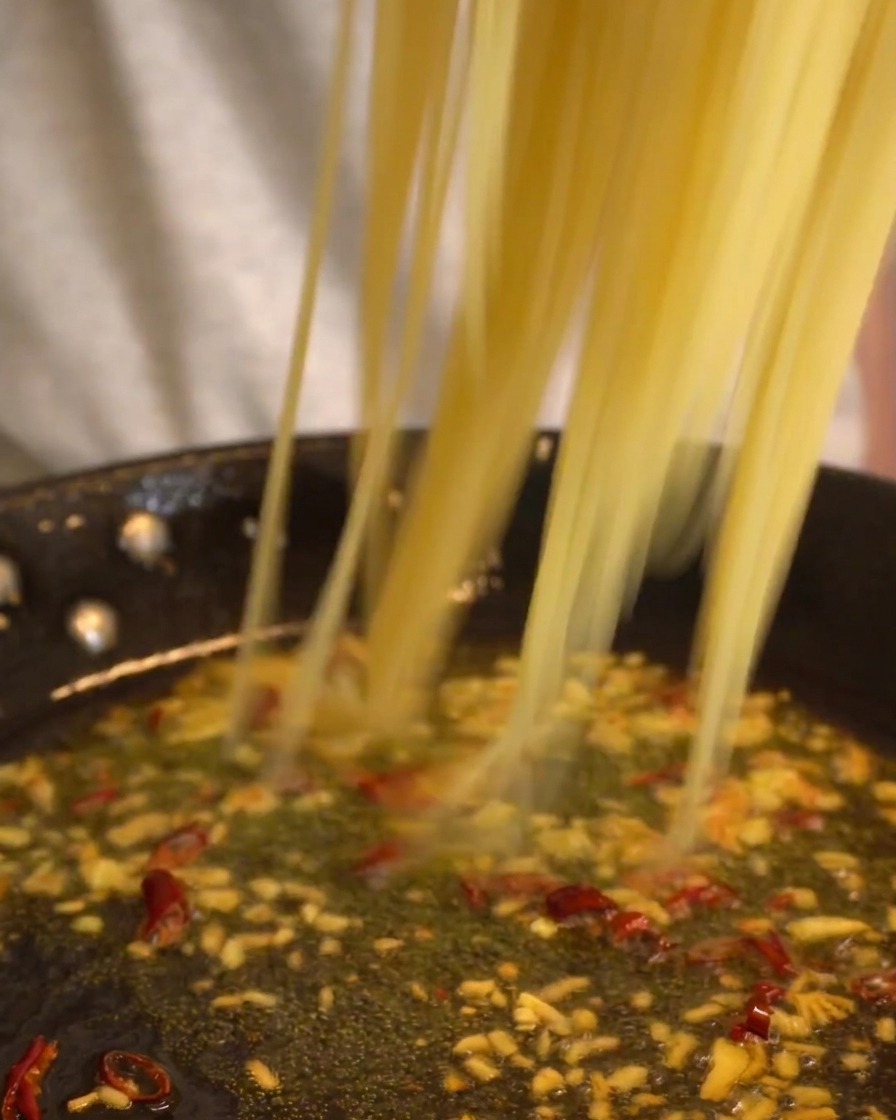 Long strands of spaghetti being dropped into a pan of simmering garlic and chili oil sauce.