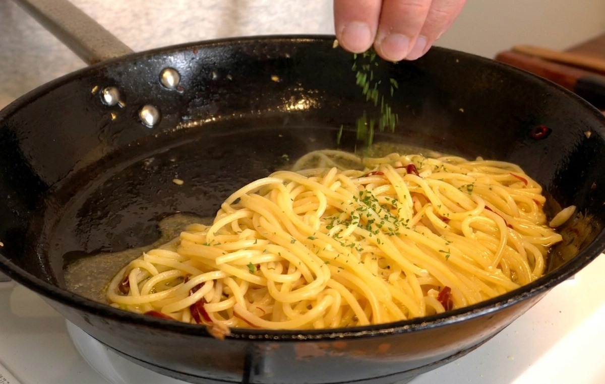 A hand sprinkling freshly chopped green parsley over a pan of glossy spaghetti.