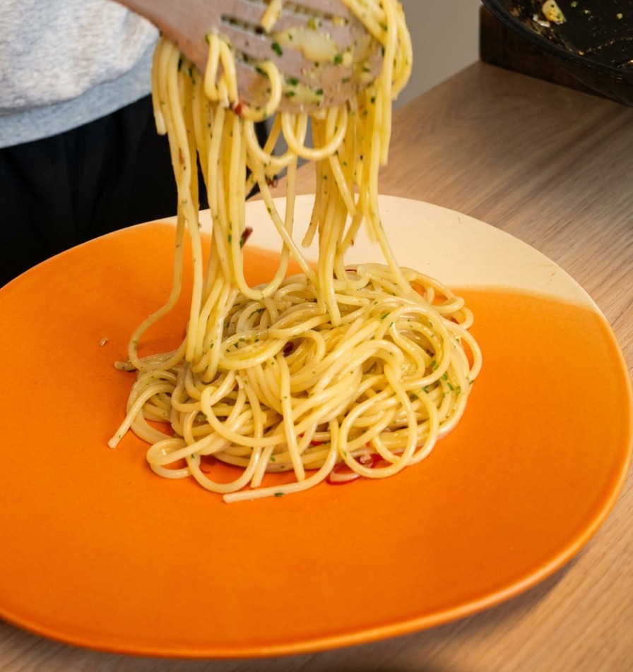 Tongs lifting and twirling spaghetti aglio e olio onto a bright orange serving plate.