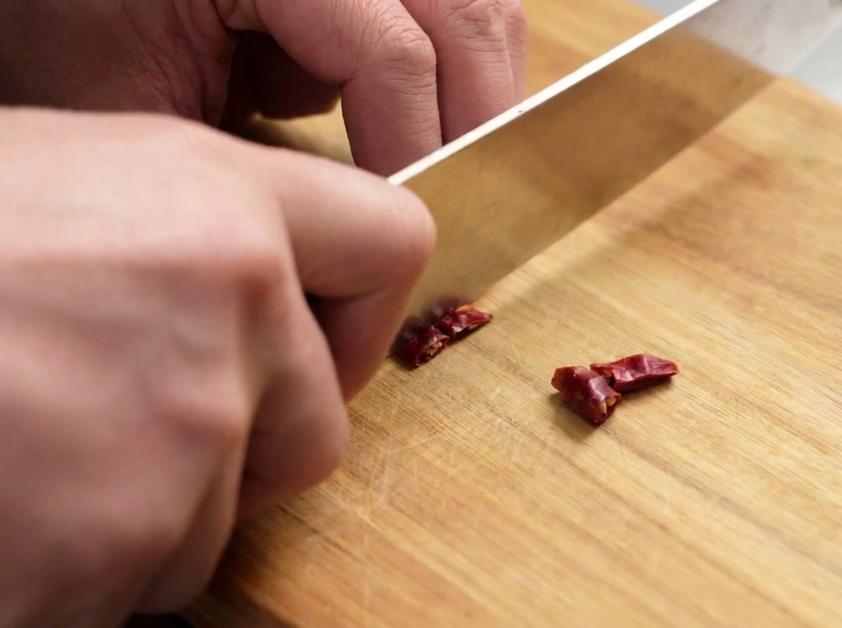 A knife cutting dried red chilies into small pieces on a wooden board.