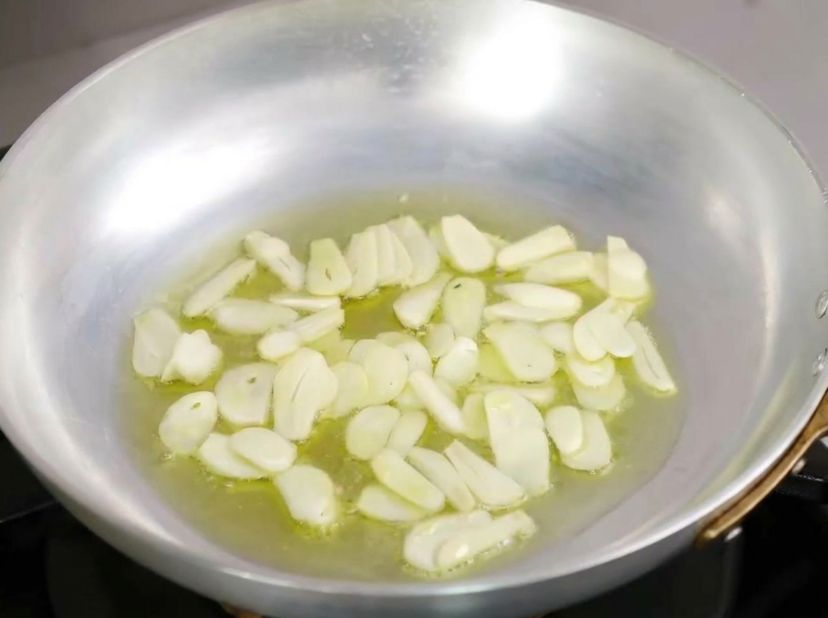 Thin garlic slices slowly sizzling in a pool of olive oil inside a metal skillet.
