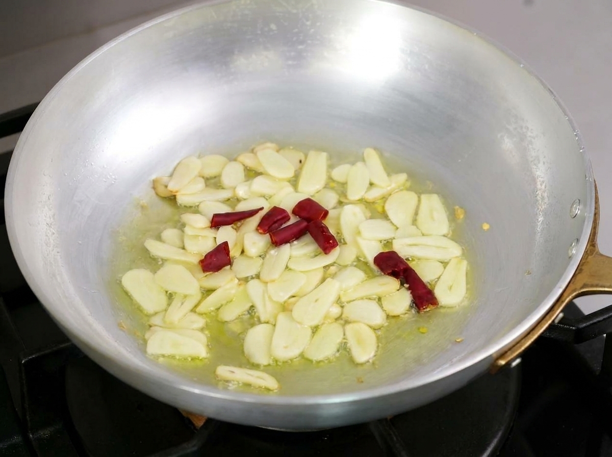 Sliced garlic and pieces of dried red chili gently sizzling in olive oil inside a metal pan.