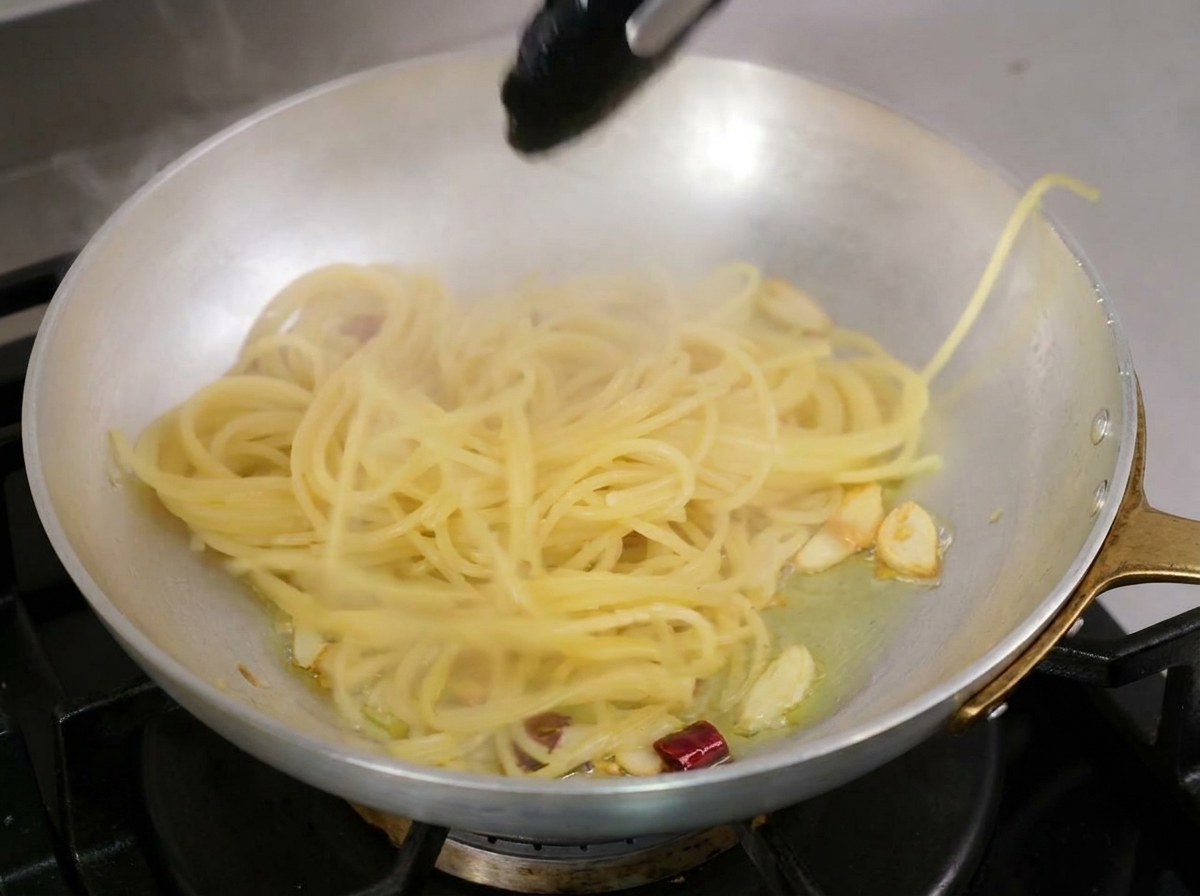 Cooked spaghetti being lifted into a pan of hot garlic and chili infused olive oil with black tongs.