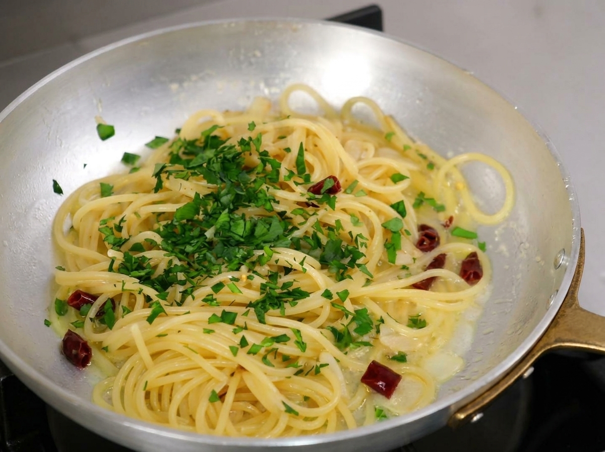 Chopped fresh parsley sprinkled generously over cooked spaghetti in a metal pan.