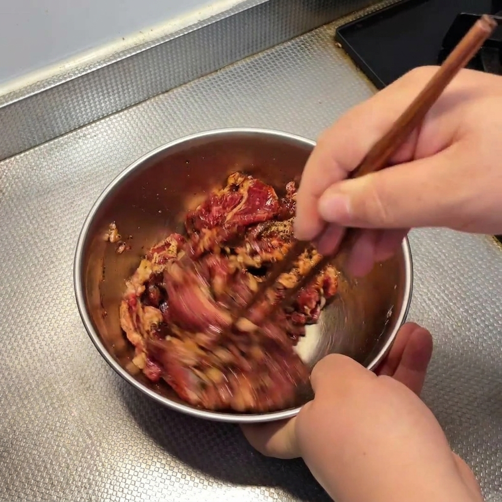 Hands using wooden chopsticks to quickly stir and marinate raw beef in a metal bowl.