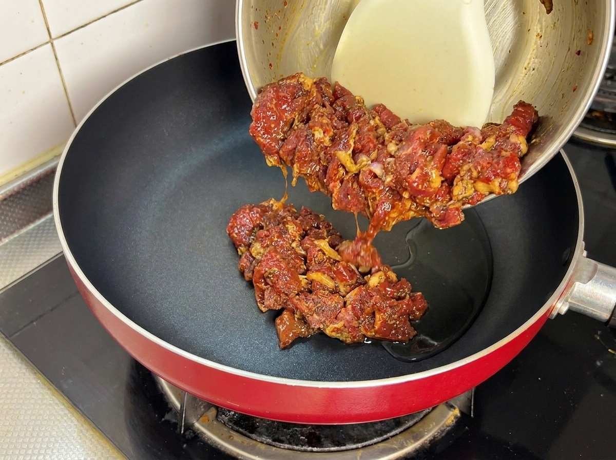 Raw marinated beef being poured from a bowl into a hot pan with cooking oil.