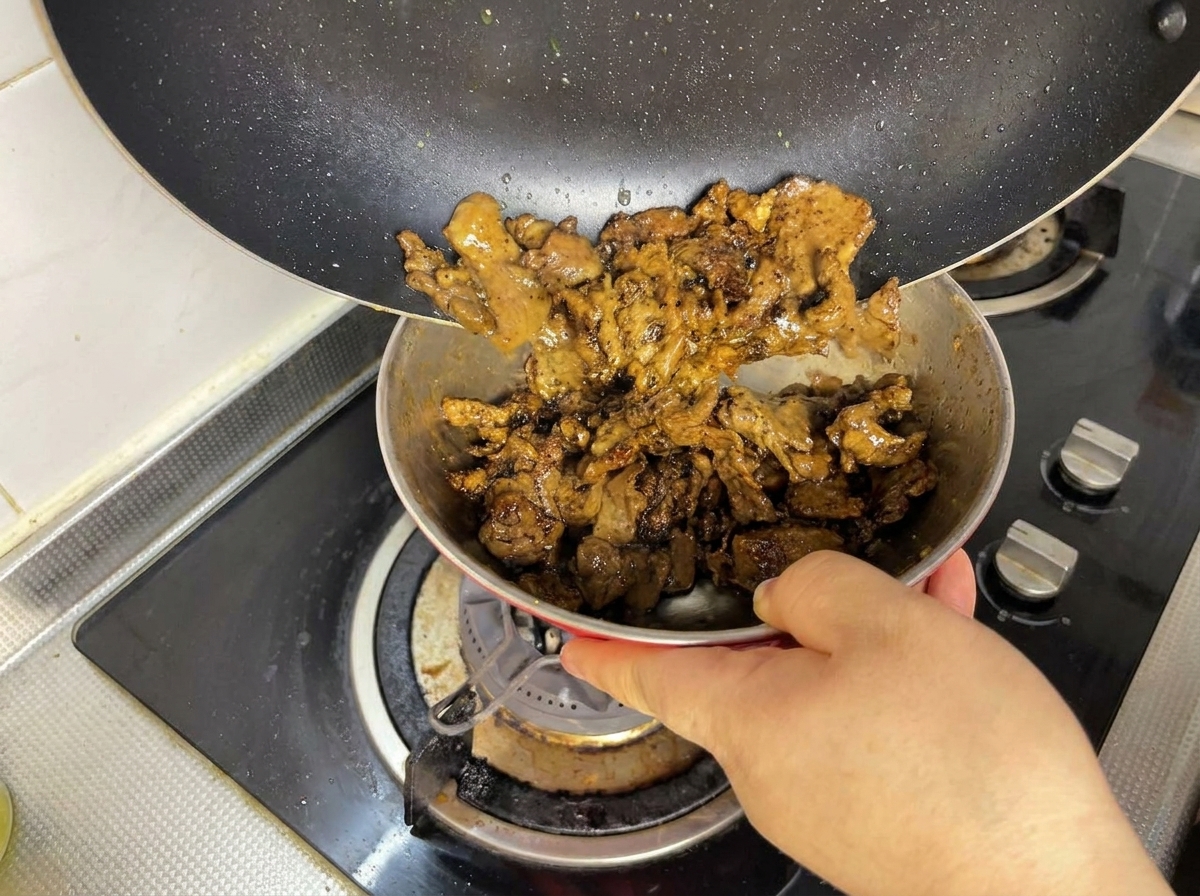 Browned pieces of beef being poured from a black frying pan into a small metal bowl.