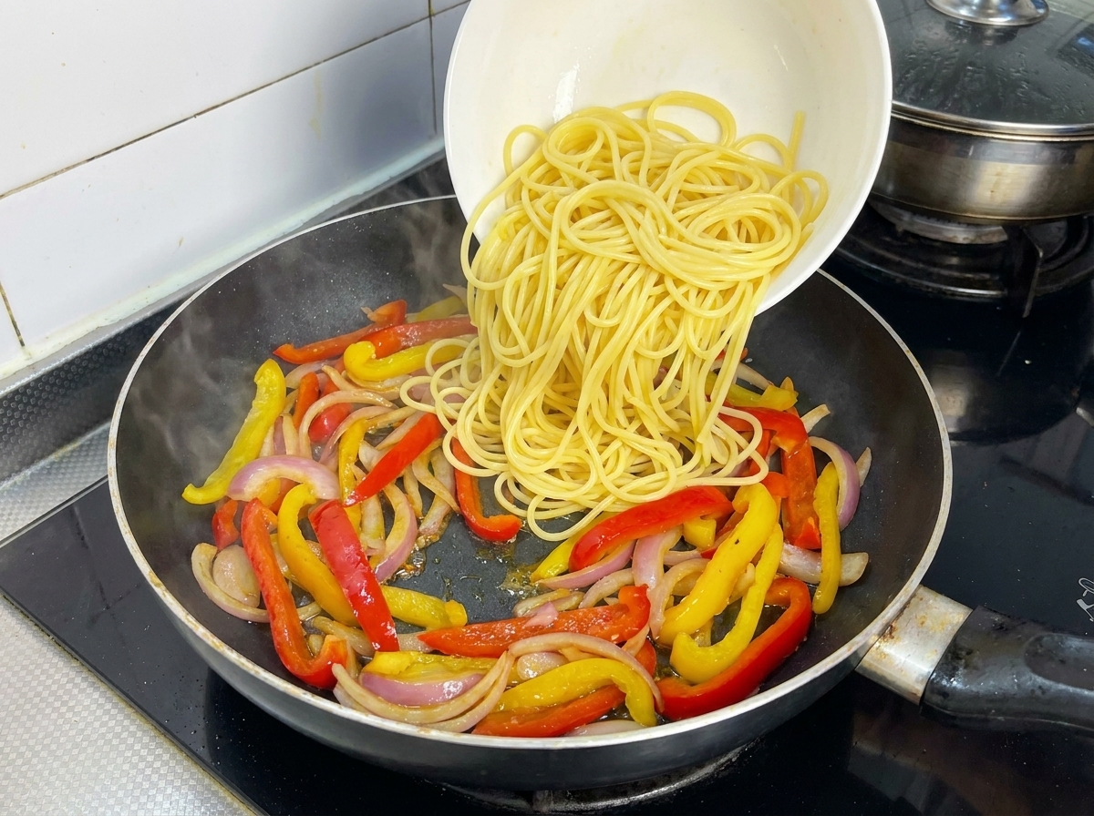 Boiled spaghetti noodles being poured from a white bowl over a mix of stir-fried bell peppers and onions in a frying pan.