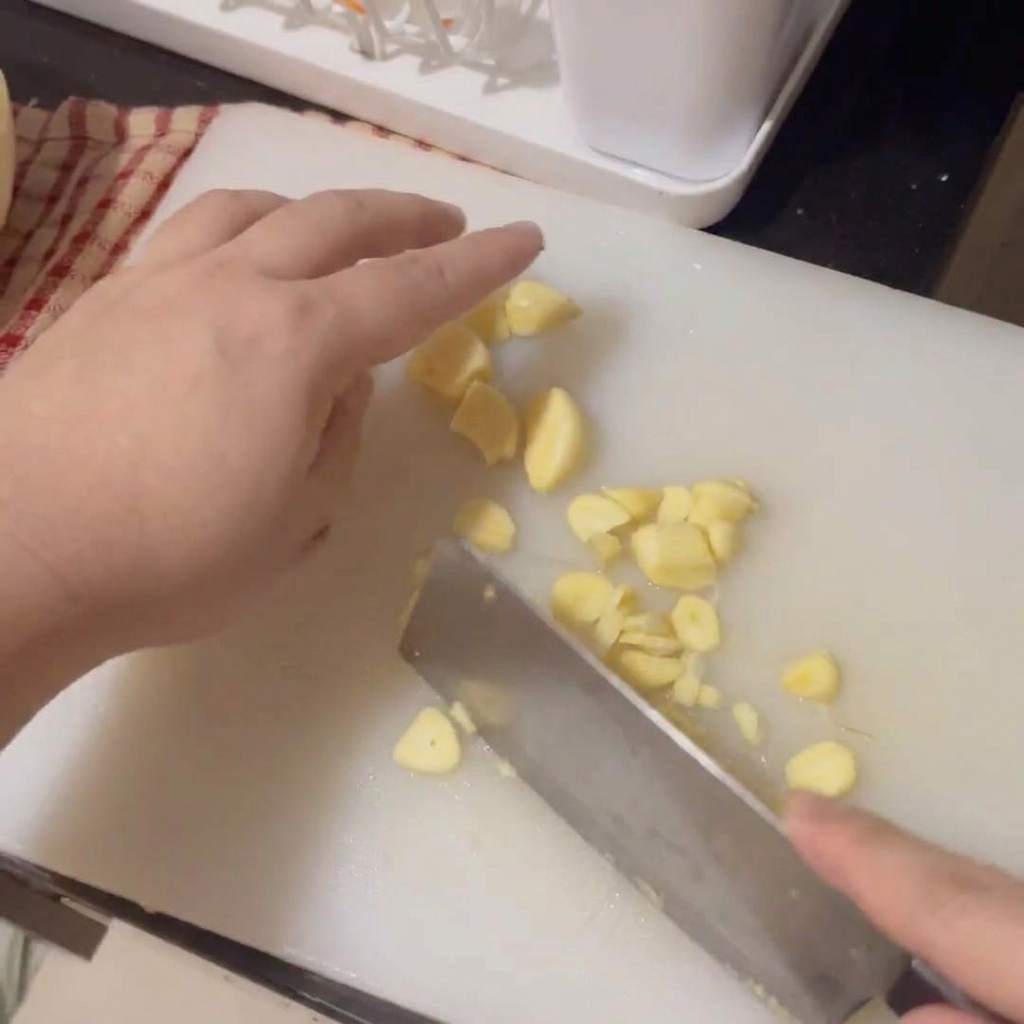 Hands using a cleaver to thinly slice fresh garlic cloves on a white cutting board.