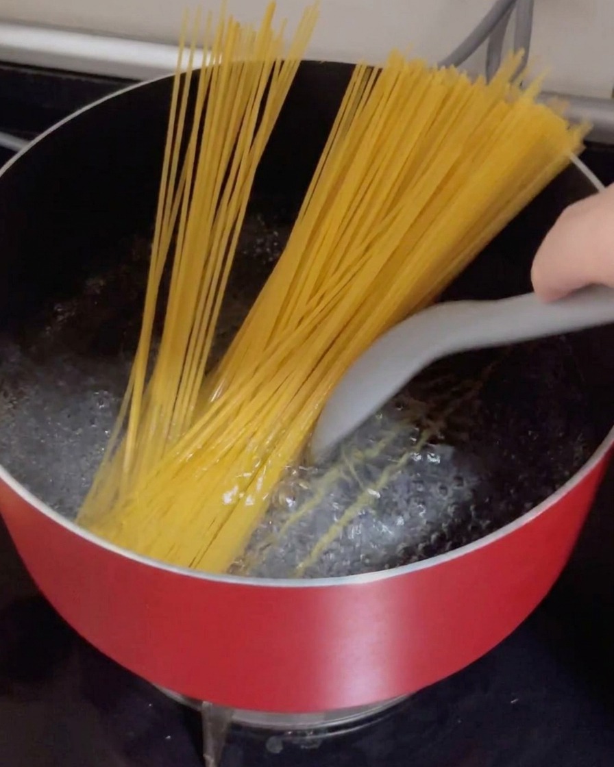 Dry spaghetti strands being pushed into a red pot of vigorously boiling water.