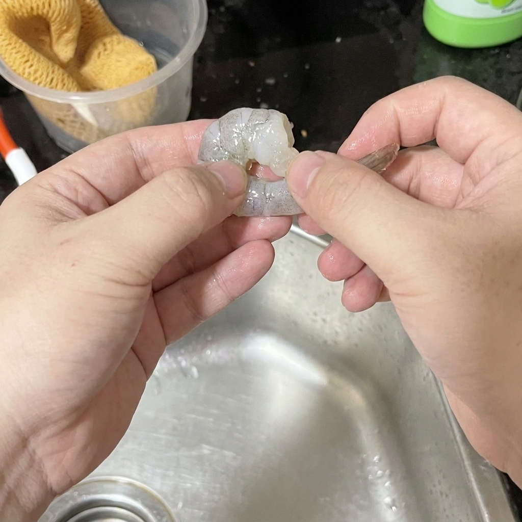 Two hands carefully peeling a raw grey shrimp over a stainless steel kitchen sink.
