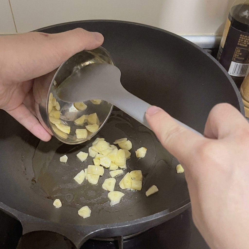 Thinly sliced garlic being scooped from a small metal bowl into a heated pan with oil.