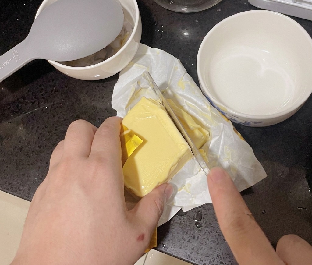 A person cutting a thick slice from a block of yellow butter using a large kitchen knife.