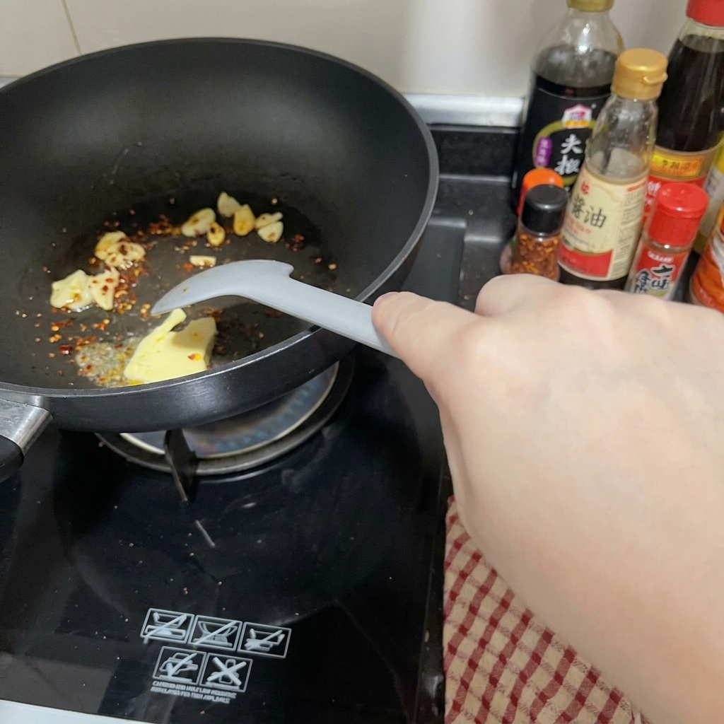 A chunk of butter melting in a black pan alongside sliced garlic and red chili flakes, being stirred with a light gray spatula.