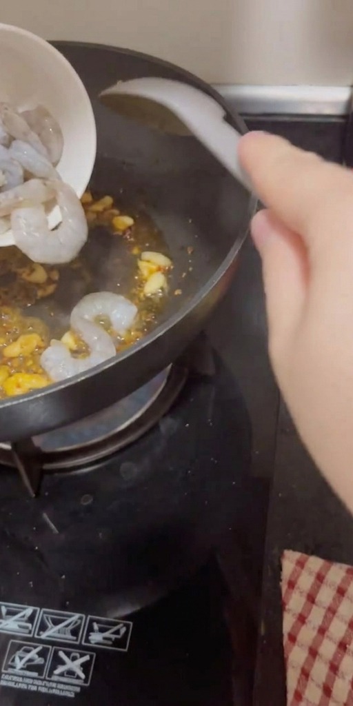 Raw, peeled shrimp being poured from a small bowl into a hot pan containing sautéed garlic and chili flakes.