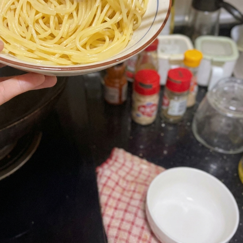 A hand holding a bowl of cooked spaghetti over a black stovetop, preparing to add it to a pan.